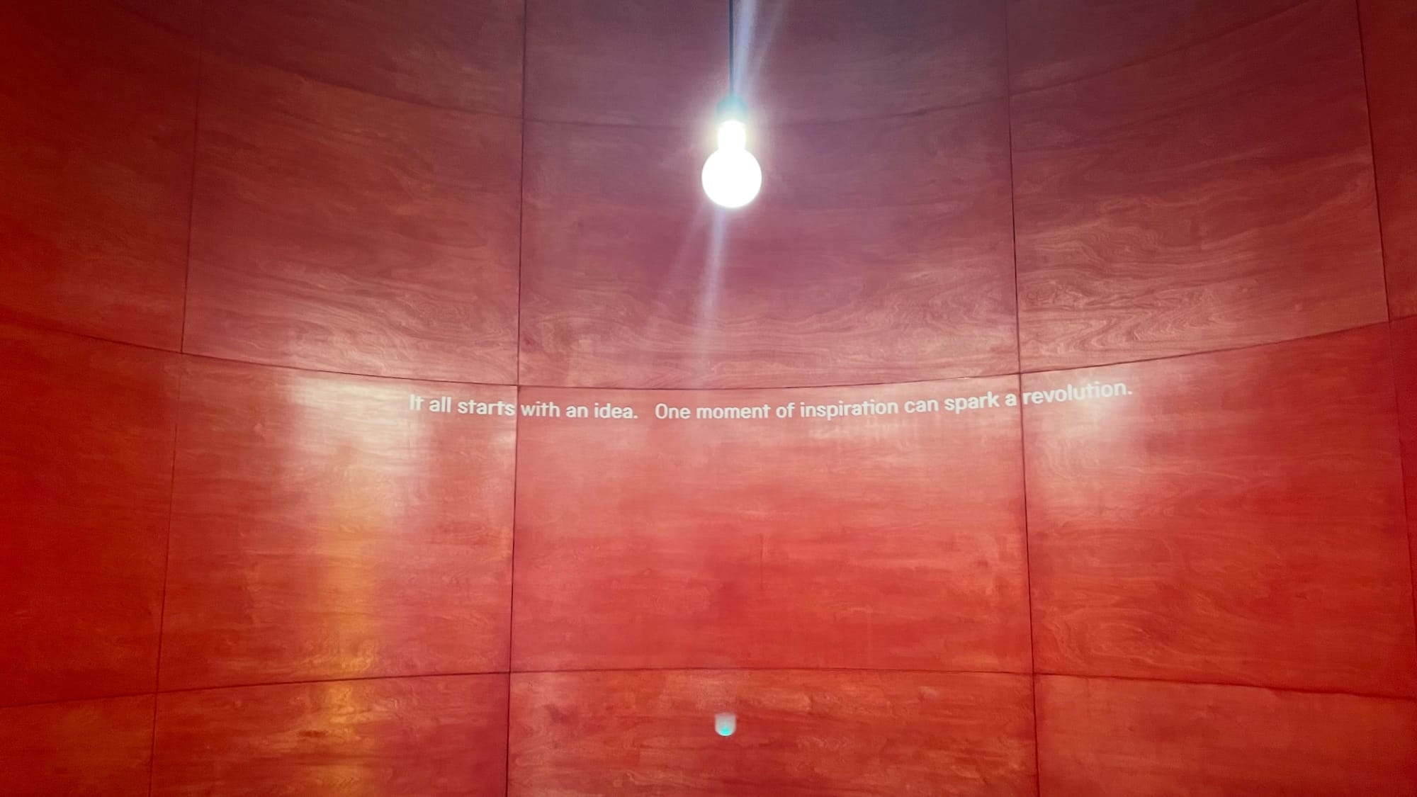 A quote is written on a red wall with a bright lightbulb hanging from the ceiling. "It all starts with an idea. One moment of inspiration can spark a revolution".