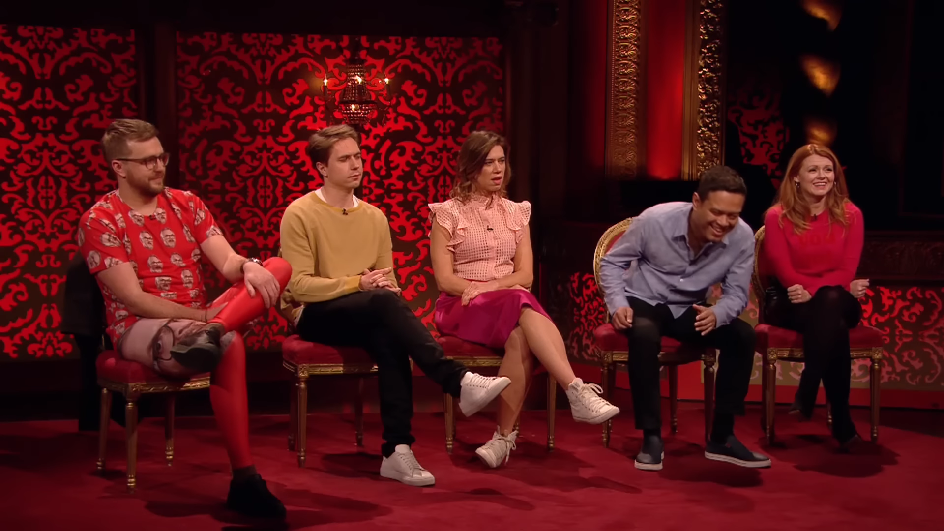 The five contestants of Taskmaster Series 8 sitting in the studio. Iain is wearing a shirt and pants both covered with pictures of Greg Davies's face, while Paul Sinha laughs in the background.
