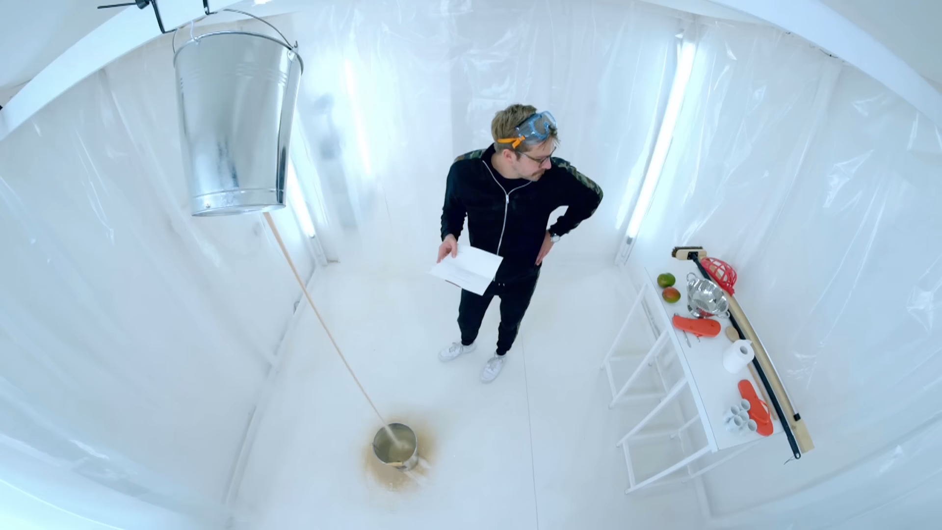 Iain Sterling standing in the Taskmaster lab, with a bucket which he moved below a bucket dripping sand. 