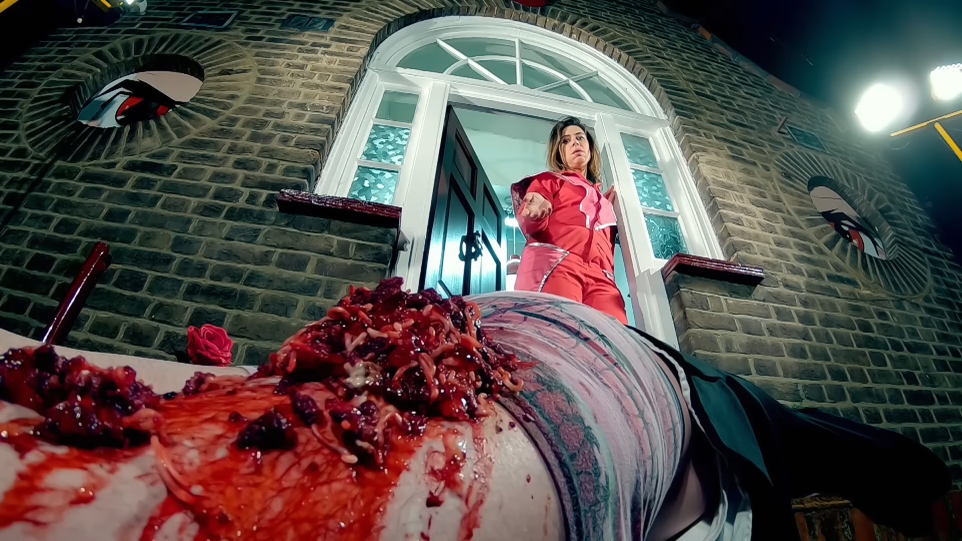 Lou Sanders in the entrance to the Taskmaster house, with Alex Horne lying on the floor with fake gore made of noodles and tomato sauce on his knee.