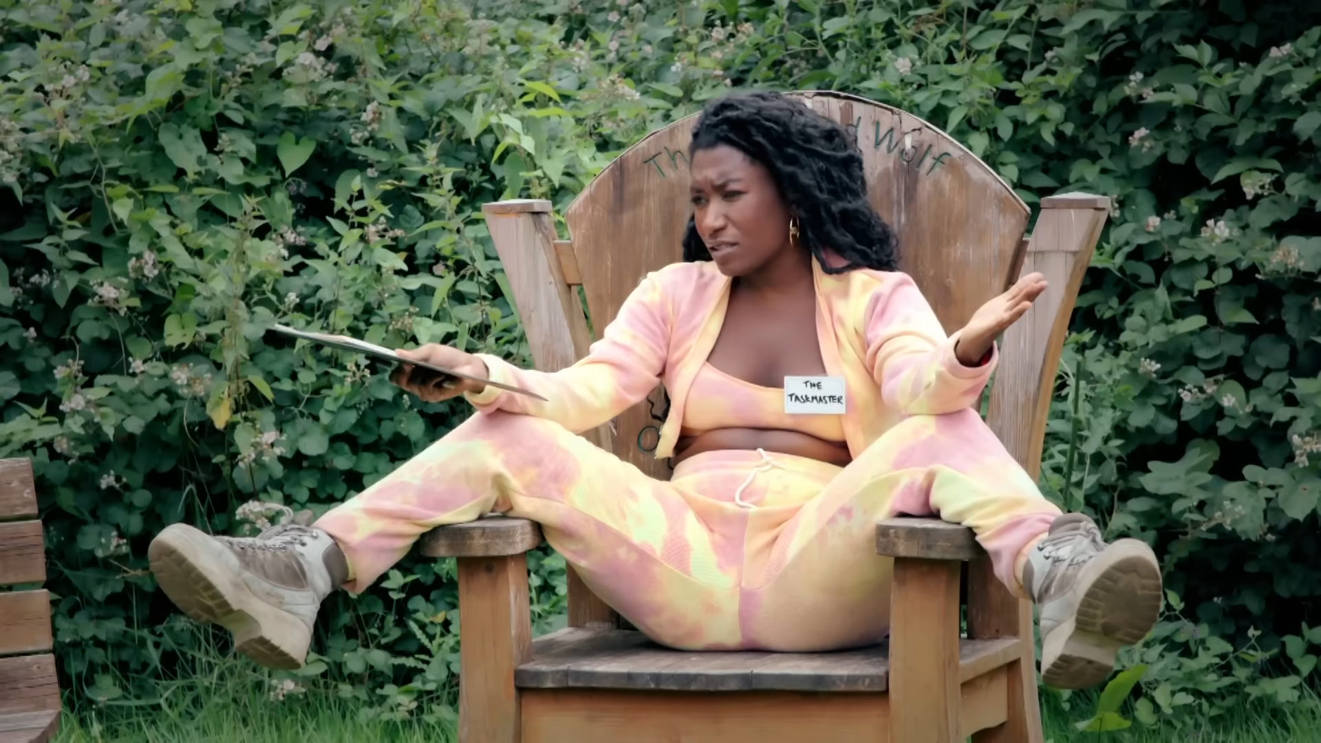 Screenshot of Taskmaster UK S13 E6. Sophie Duker pretends to be the Taskmaster, sitting on the main chair with her legs spread.